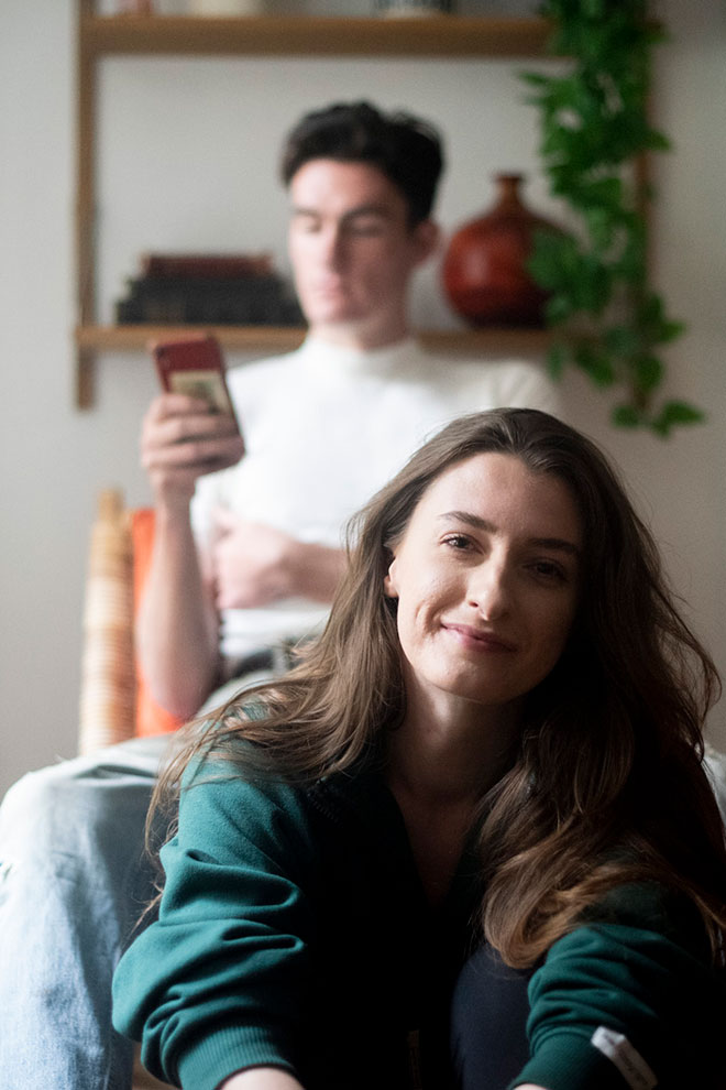 Relaxed couple on sofa, woman smiling at camera while man looks at phone, natural home setting
