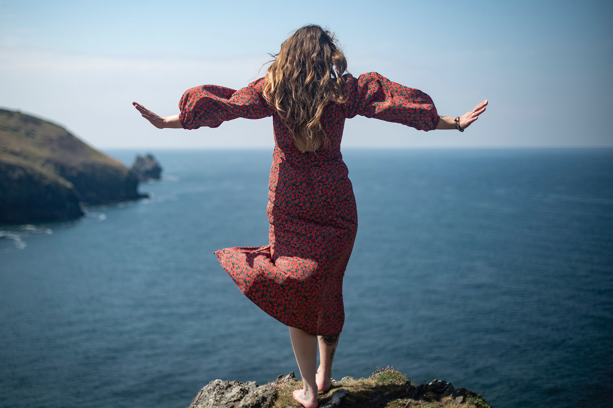Woman in red dress standing on cliff edge with arms wide, dramatic seaside portrait
