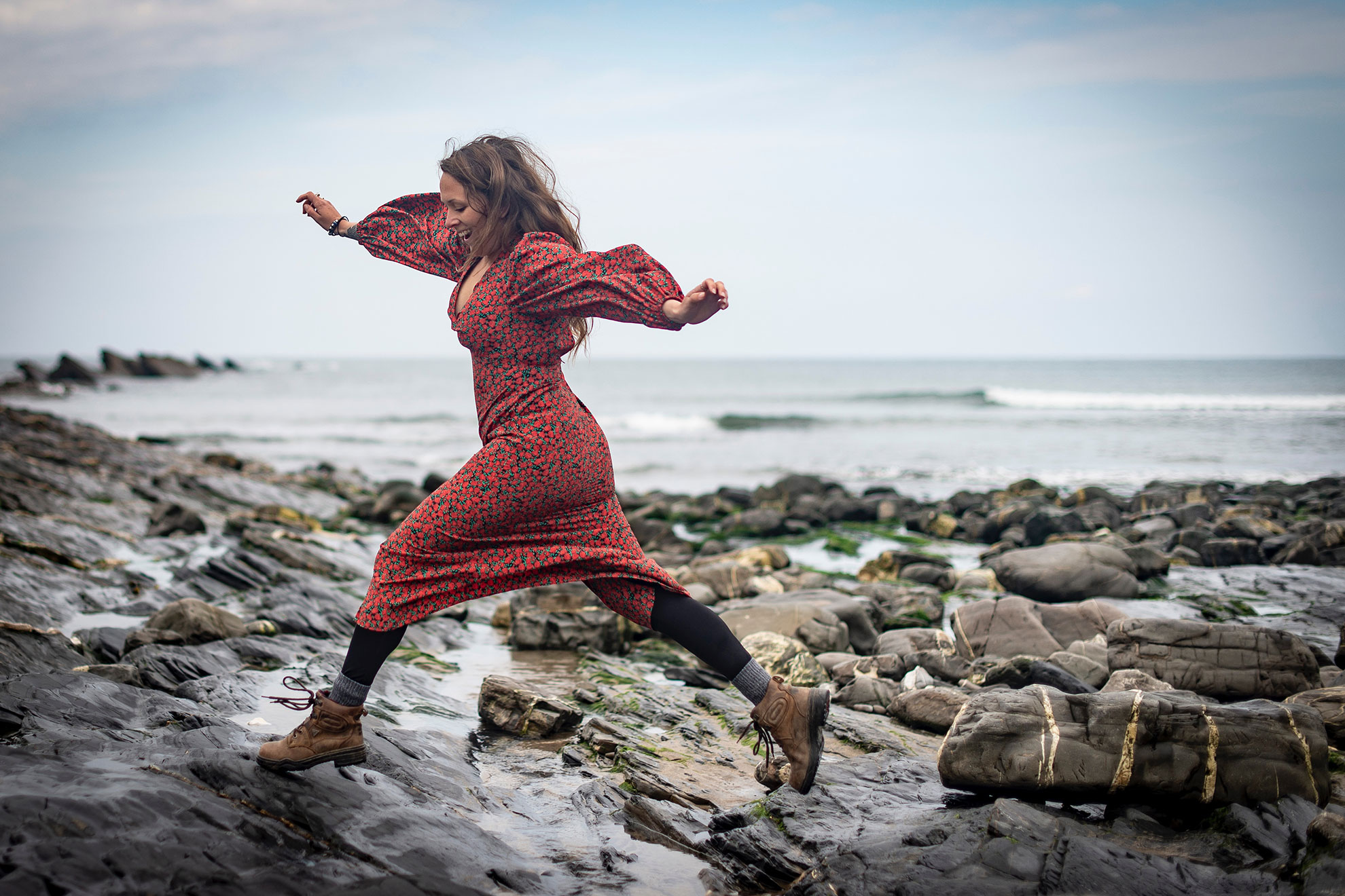 Woman in red patterned dress leaping across rocks by the sea, coastal fashion photography