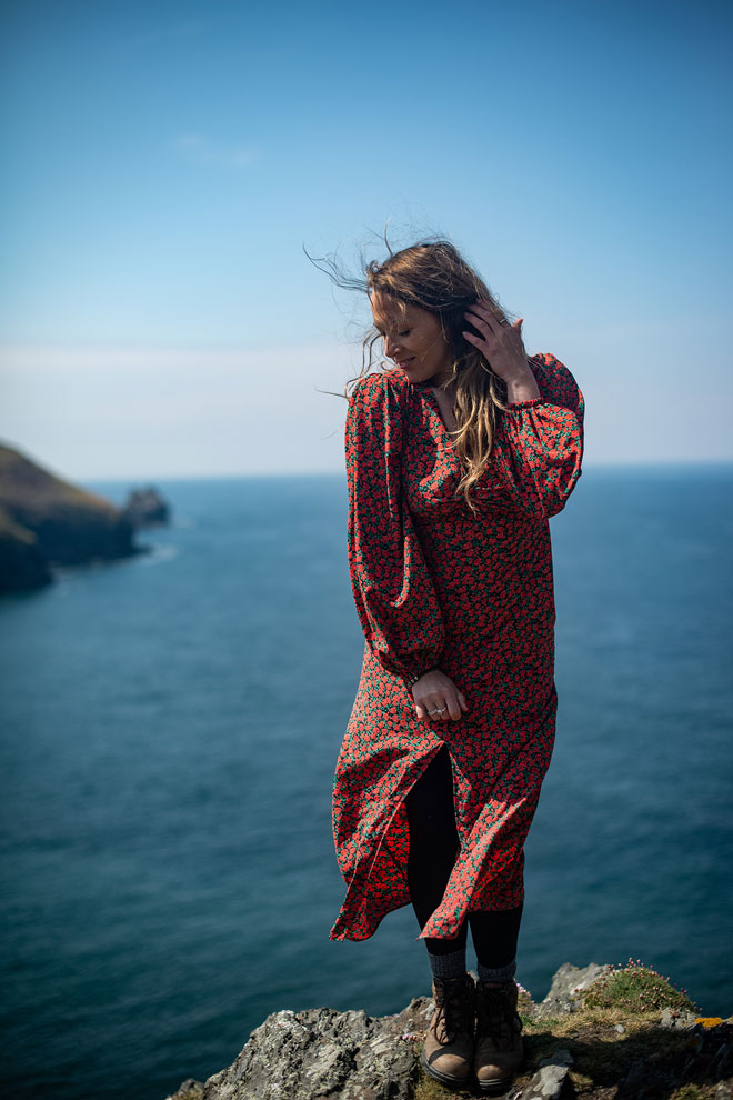 Model in red dress posing on clifftop with blue ocean in background, lifestyle fashion shoot
