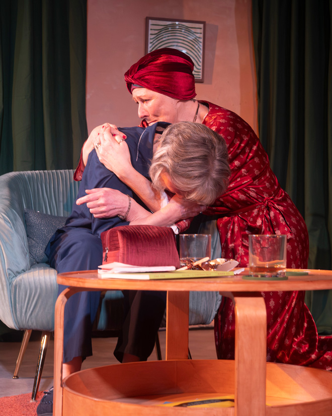 Woman in red embraces the grey-haired woman in blue, who leans forward emotionally on a sofa.