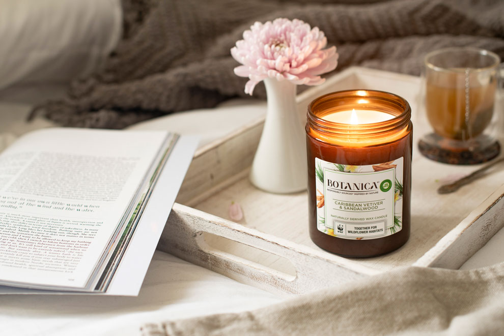 Botanica candle on a tray beside a cup of tea, pink flowers and an open book.
