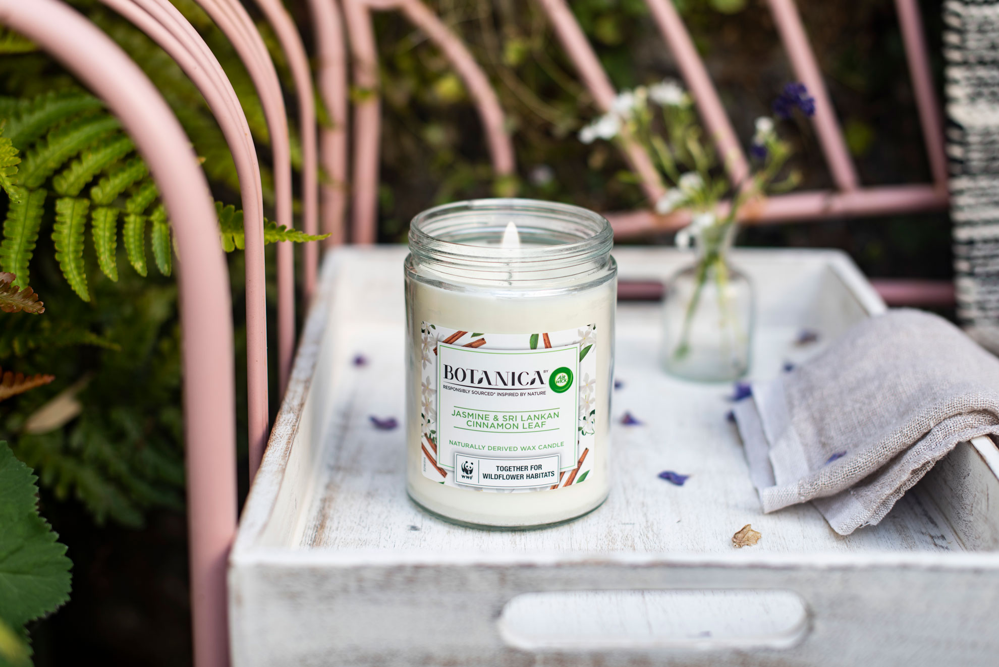 Botanica scented candle displayed in a garden setting on a wrought iron pastel pink bench.