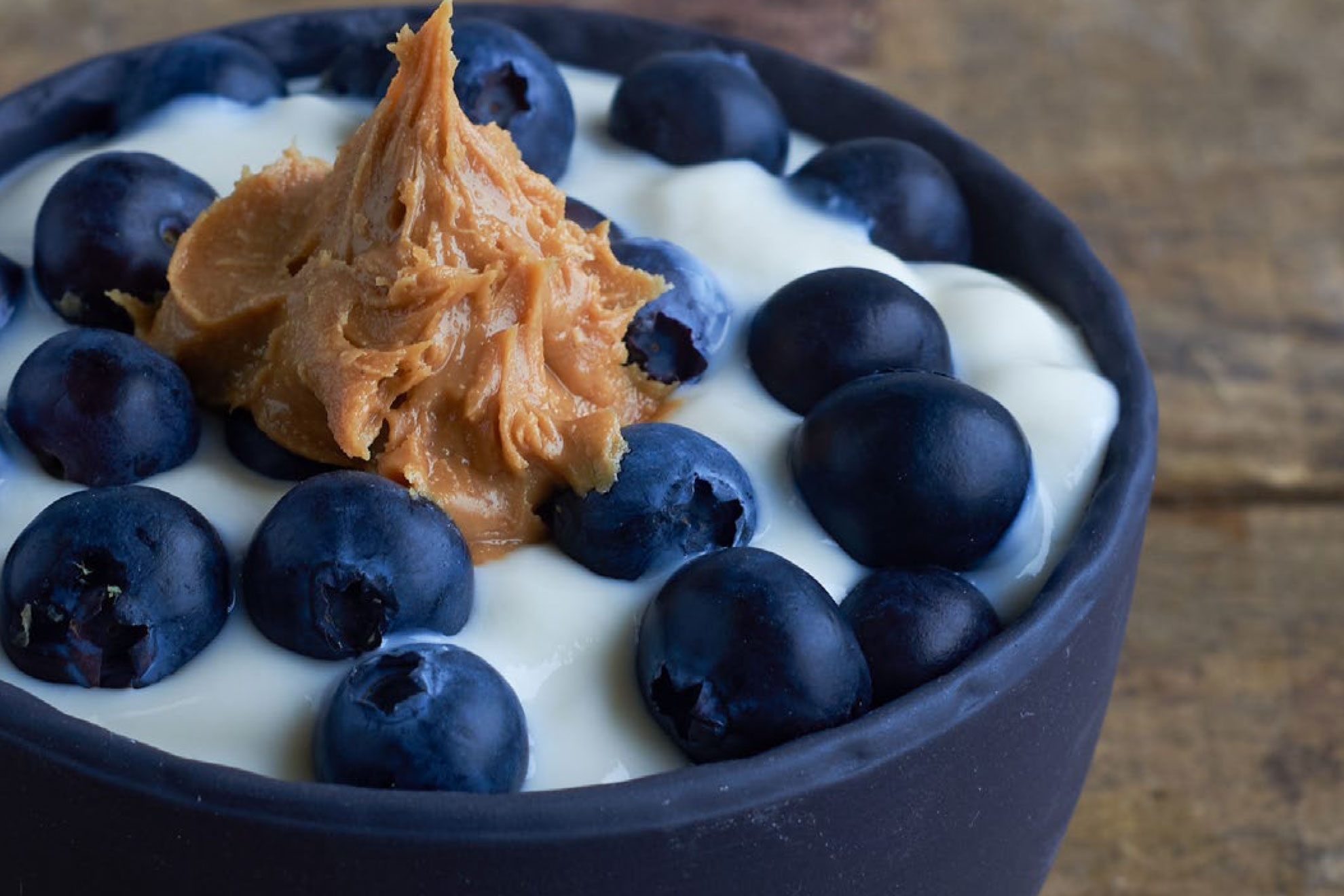 Natural yoghurt topped with fresh blueberries and peanut butter, styled in a black bowl
