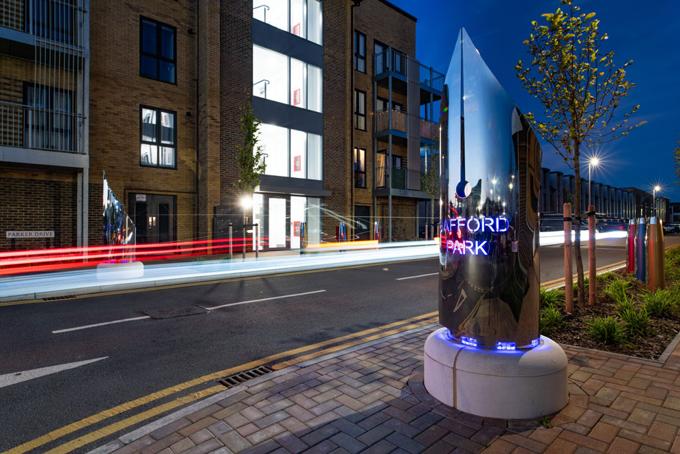 Entrance to Safford Park housing development at night, with light streaks from cars and a striking illuminated chrome sign in the shape of a pen nib